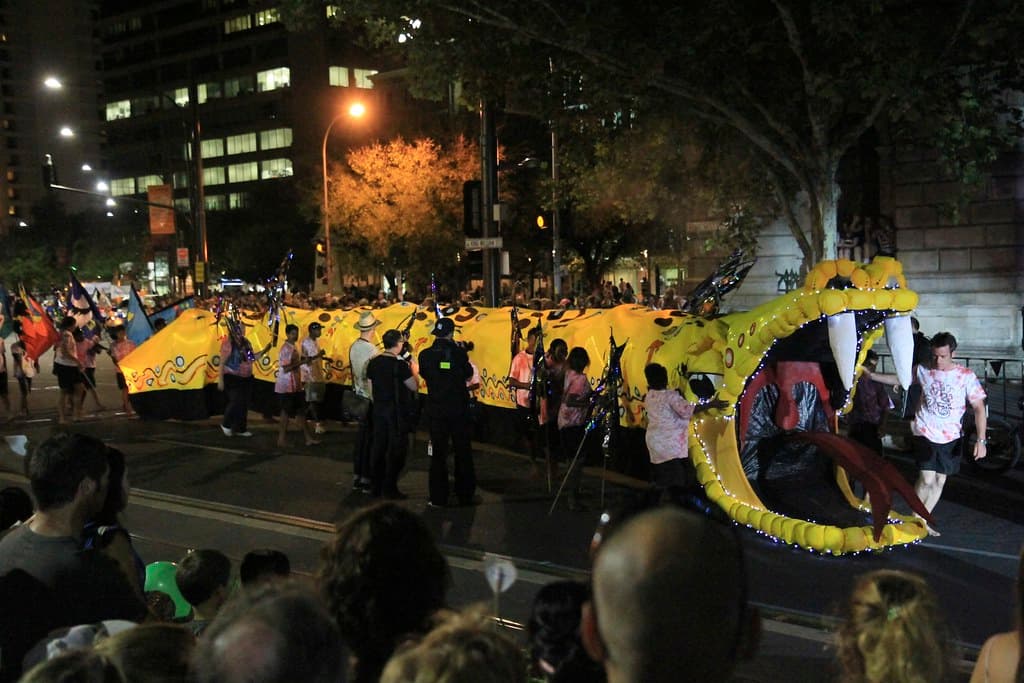 A giant golden dragon puppet is carried through a crowded city street at night during the Adelaide Fringe parade