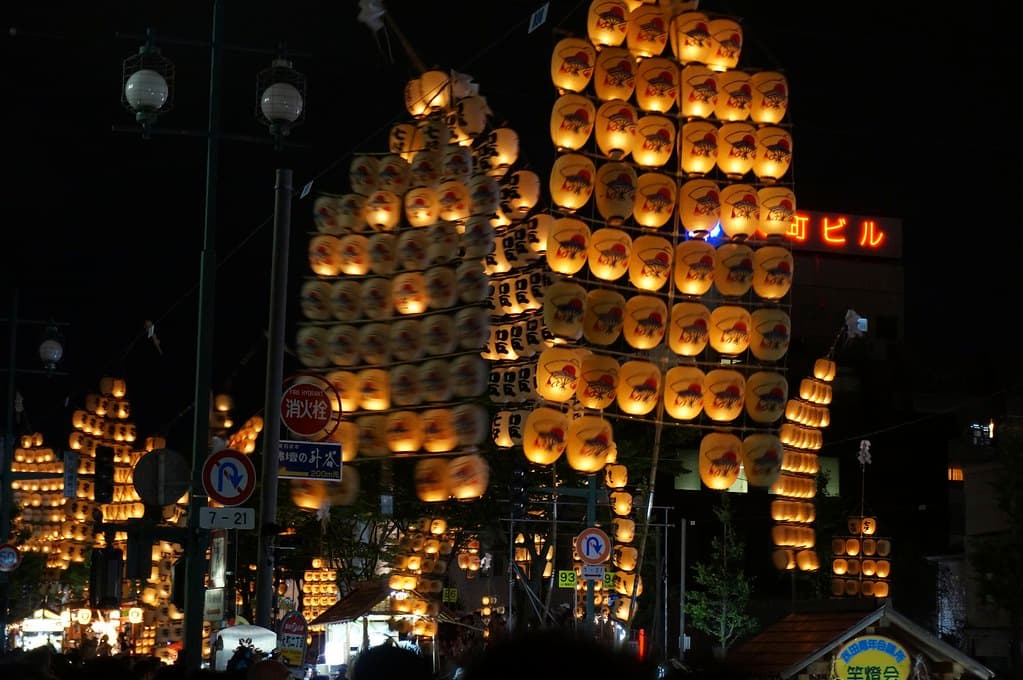 Tall bamboo kantō poles hung with dozens of glowing paper lanterns sway above a night crowd in Akita