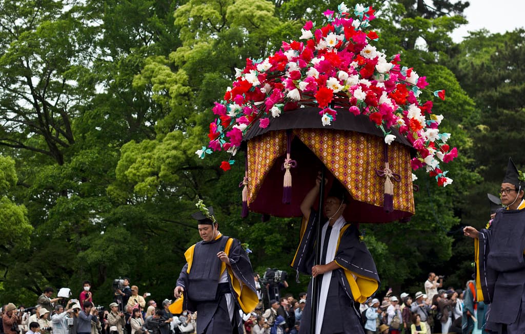 A large parasol topped with flowers is carried in the Aoi Matsuri procession past crowds under green trees in Kyoto