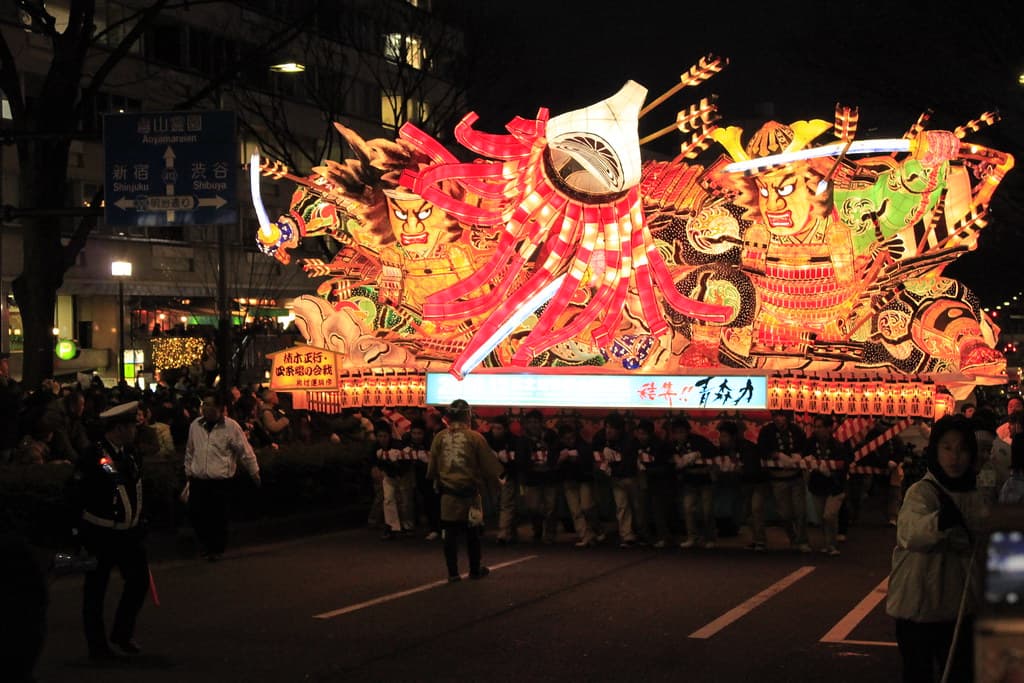 A giant illuminated Nebuta warrior float glowing at night as it moves down an Aomori street