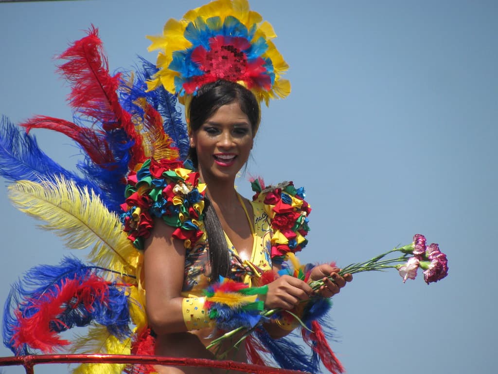 A smiling woman in a feathered costume holding flowers at the Barranquilla Carnival