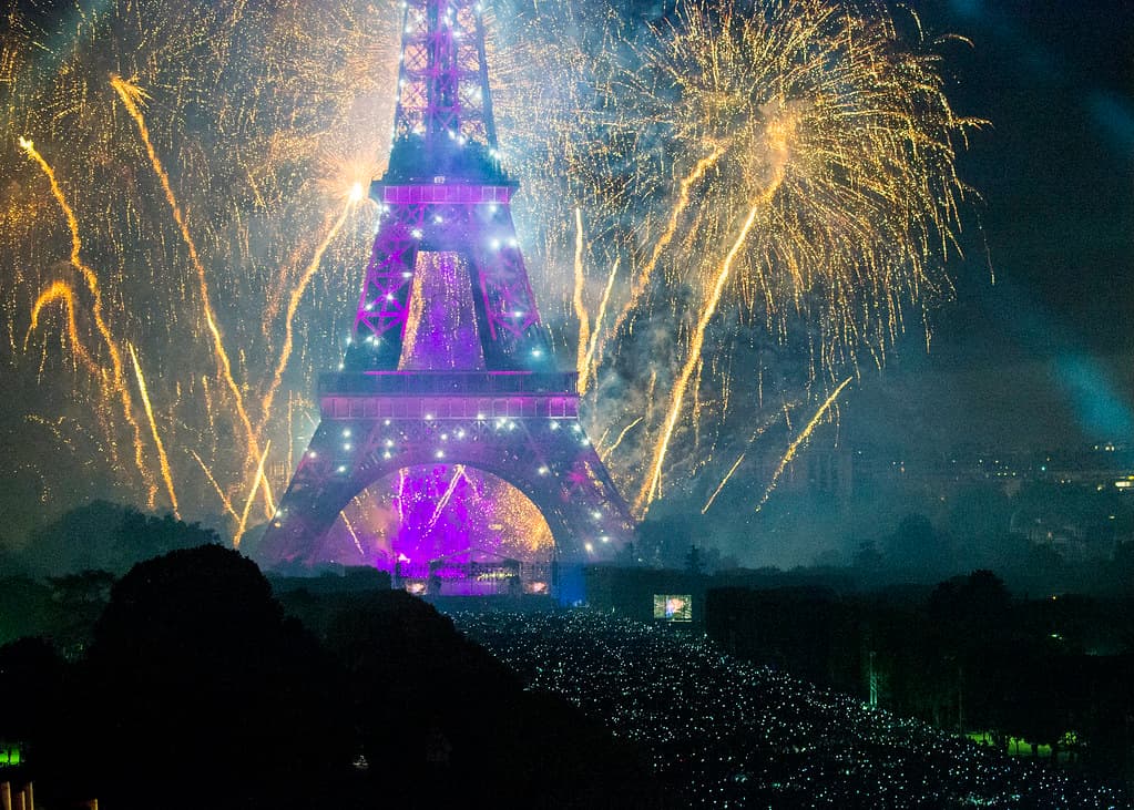 Fireworks in gold and purple burst around the illuminated Eiffel Tower on Bastille Day night