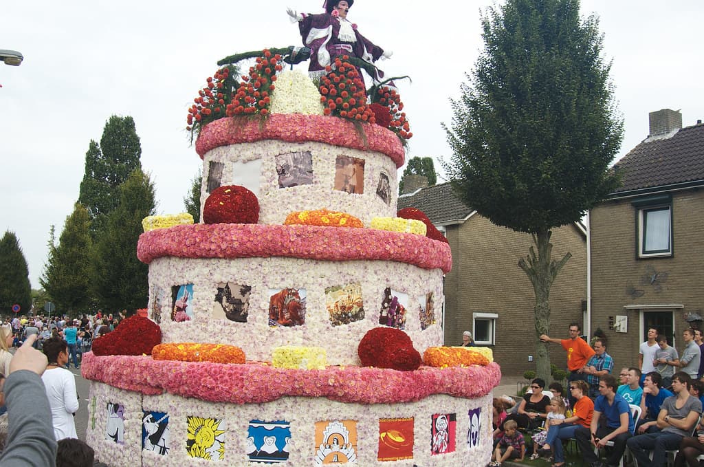 A towering float shaped like a layered cake, covered in dahlias, passes spectators on a street in Zundert