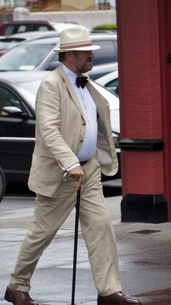 A man in a cream Edwardian suit, bow tie and panama hat walks with a cane along a Dublin street on Bloomsday