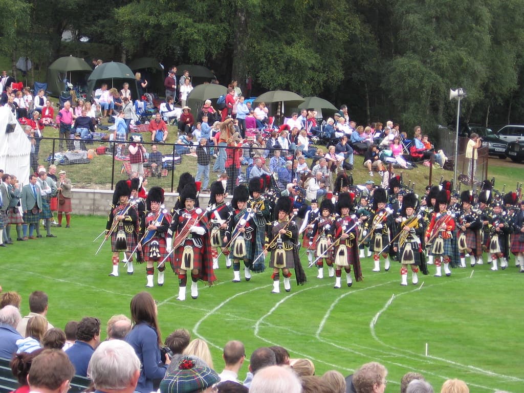 Massed pipe bands in kilts march across the green field at the Braemar Gathering, with grandstands full of spectators behind