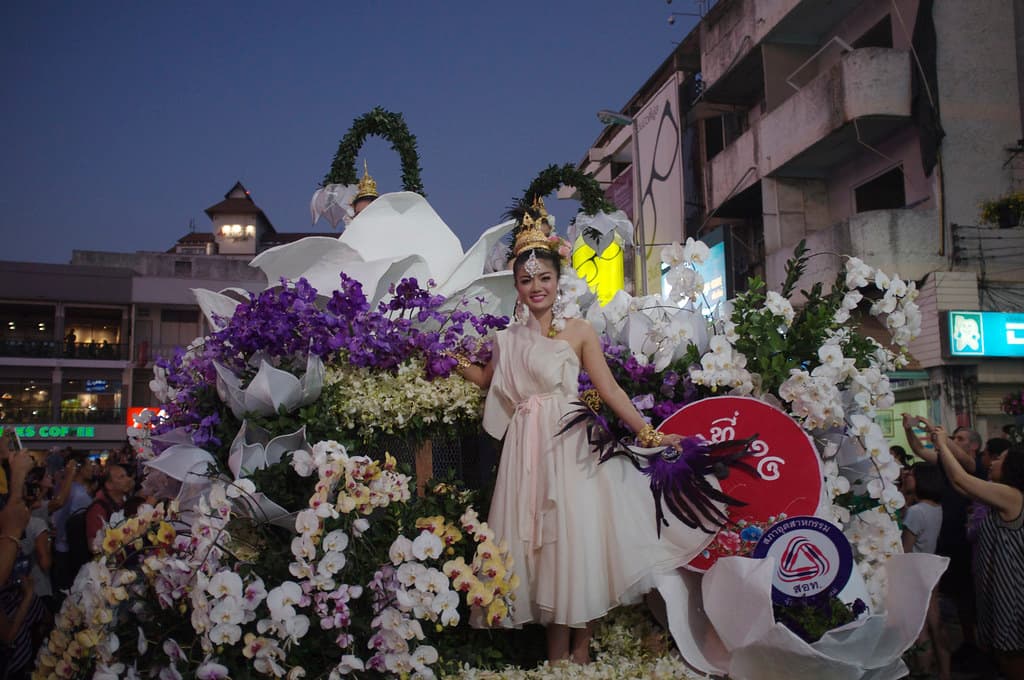 A float covered in white and purple flowers, carrying a woman in a white dress, at dusk during the Chiang Mai Flower Festival