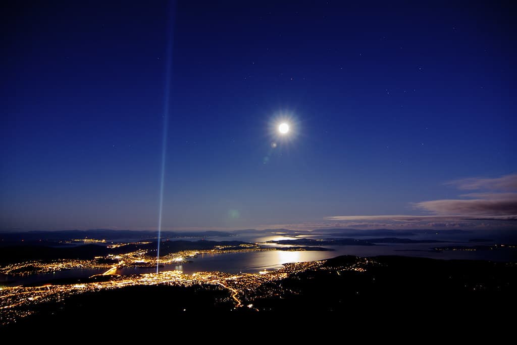 A beam of light rises from the waterfront into a full-moon sky above the lights of Hobart, seen from kunanyi/Mount Wellington