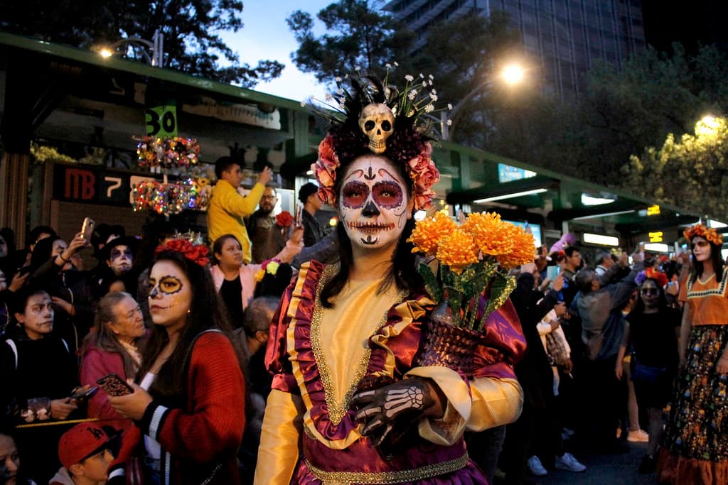 A woman dressed as La Catrina carries an armful of marigolds through a night procession in Mexico City