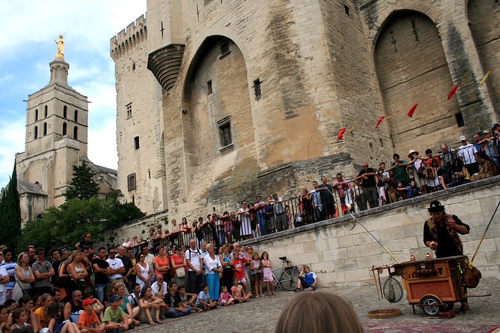 A crowd sits on the steps below the stone walls of the Palais des Papes in Avignon watching a street performer during the festival