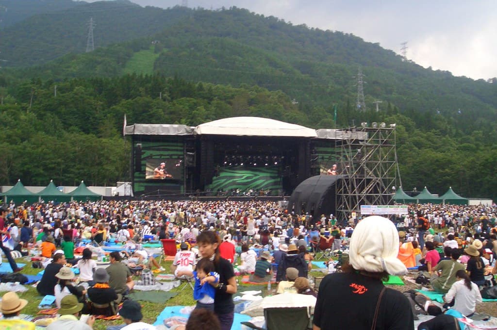 Crowds sit and stand on the grass in front of the Green Stage at Fuji Rock, with forested mountains behind