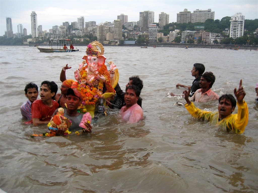 Devotees wade into the sea carrying a Ganesh idol for immersion, with a city skyline across the bay