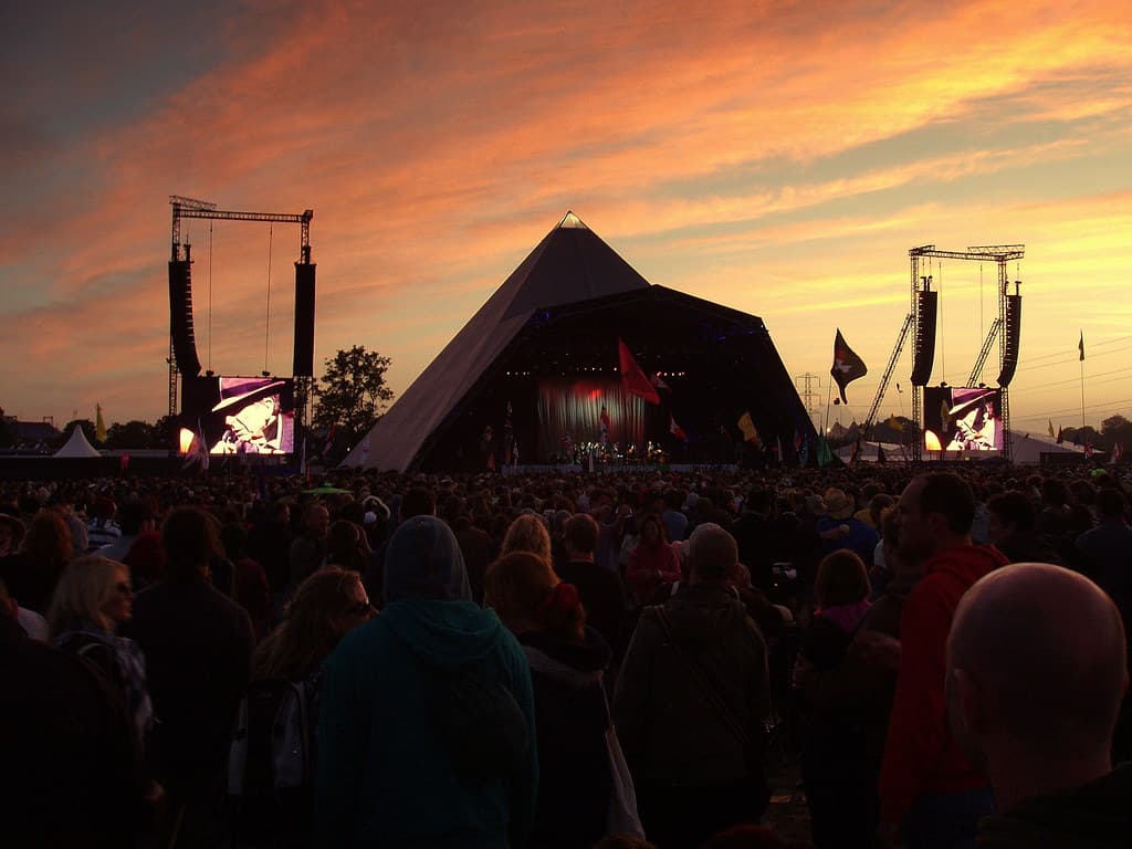 Crowds face the Pyramid Stage at Glastonbury Festival under a fiery orange sunset sky