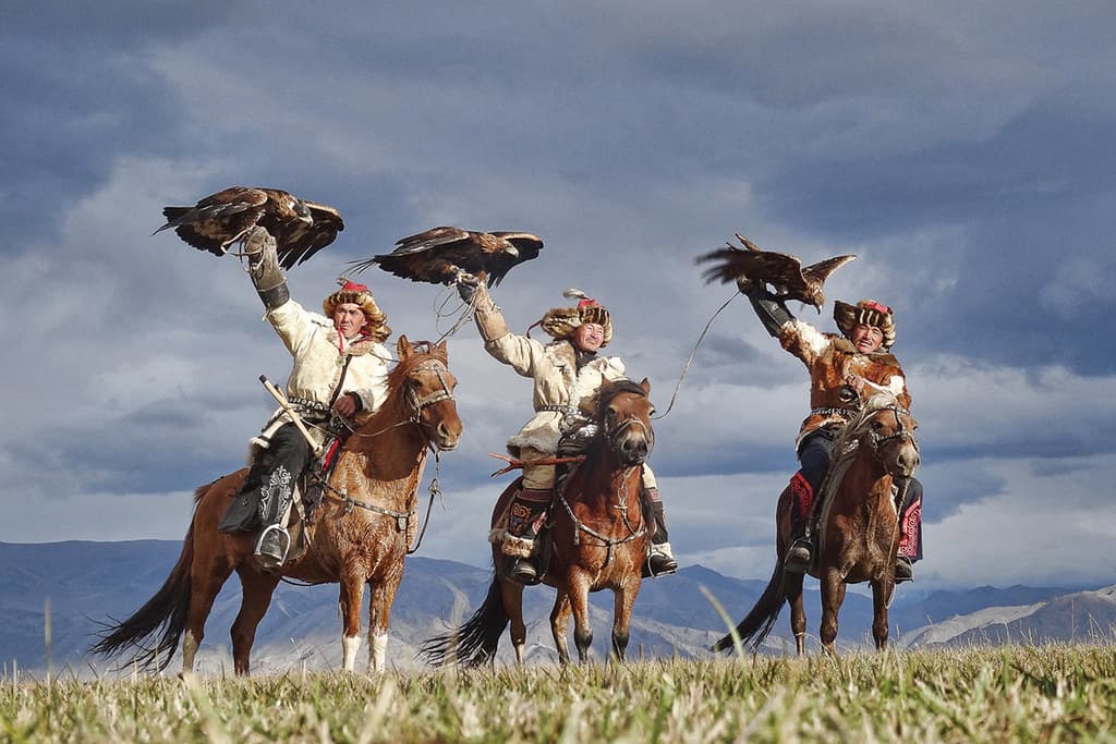 Three Kazakh eagle hunters on horseback raise golden eagles with outstretched wings against a stormy sky in western Mongolia
