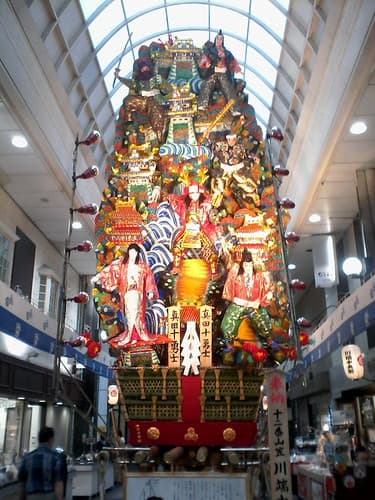 A towering decorated kazariyama float covered in warrior figures and waves stands in a covered shopping arcade in Hakata