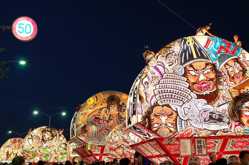 Illuminated neputa floats with painted warriors parade through a Hirosaki street at dusk