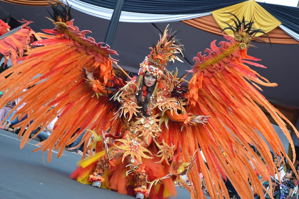 A performer in a vast flame-orange feathered costume spreads its wings on the street catwalk at Jember Fashion Carnaval