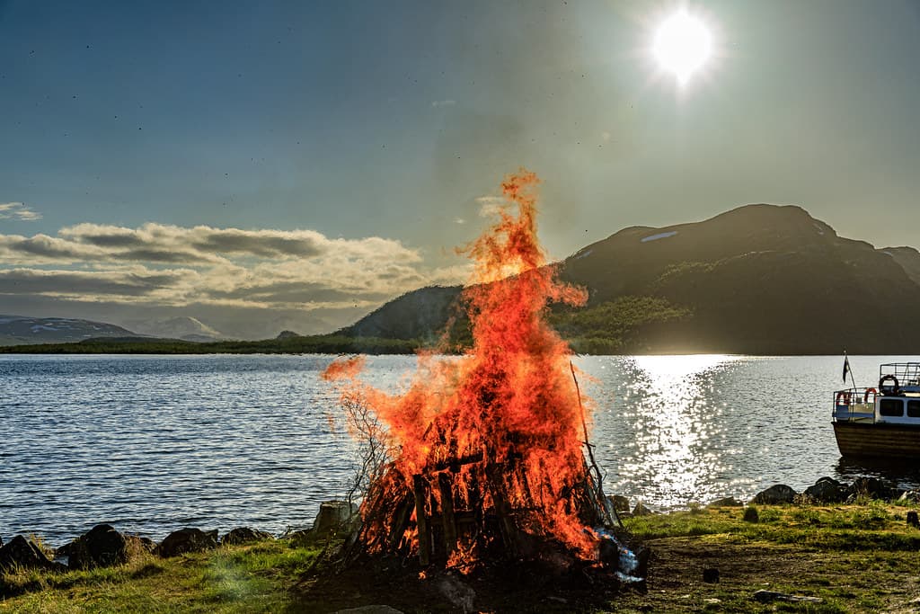 A midsummer bonfire blazes on a lakeshore under the evening sun, with fells across the water in Kilpisjärvi, Finnish Lapland