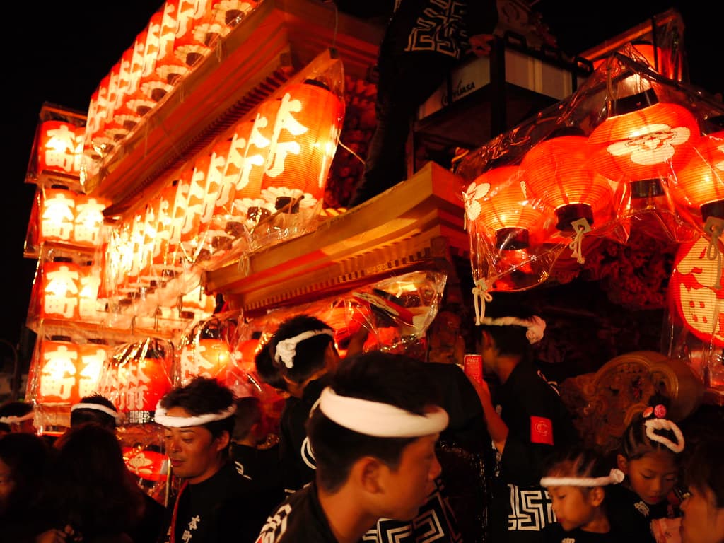 Men in happi coats and headbands crowd around a danjiri float hung with glowing red lanterns at night in Kishiwada