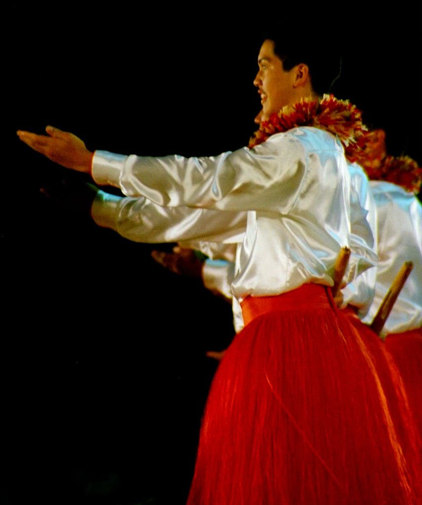 Male hula dancers in white satin shirts, red skirts and red leis perform with pūʻili sticks at the Merrie Monarch Festival in Hilo