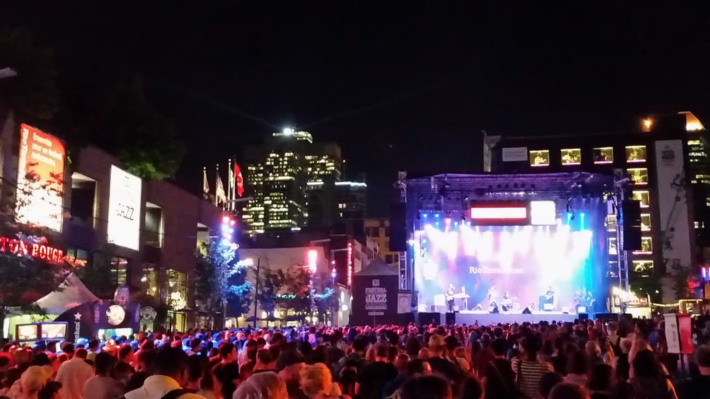 A huge crowd fills a downtown square at night in front of a brightly lit outdoor stage at the Montreal International Jazz Festival