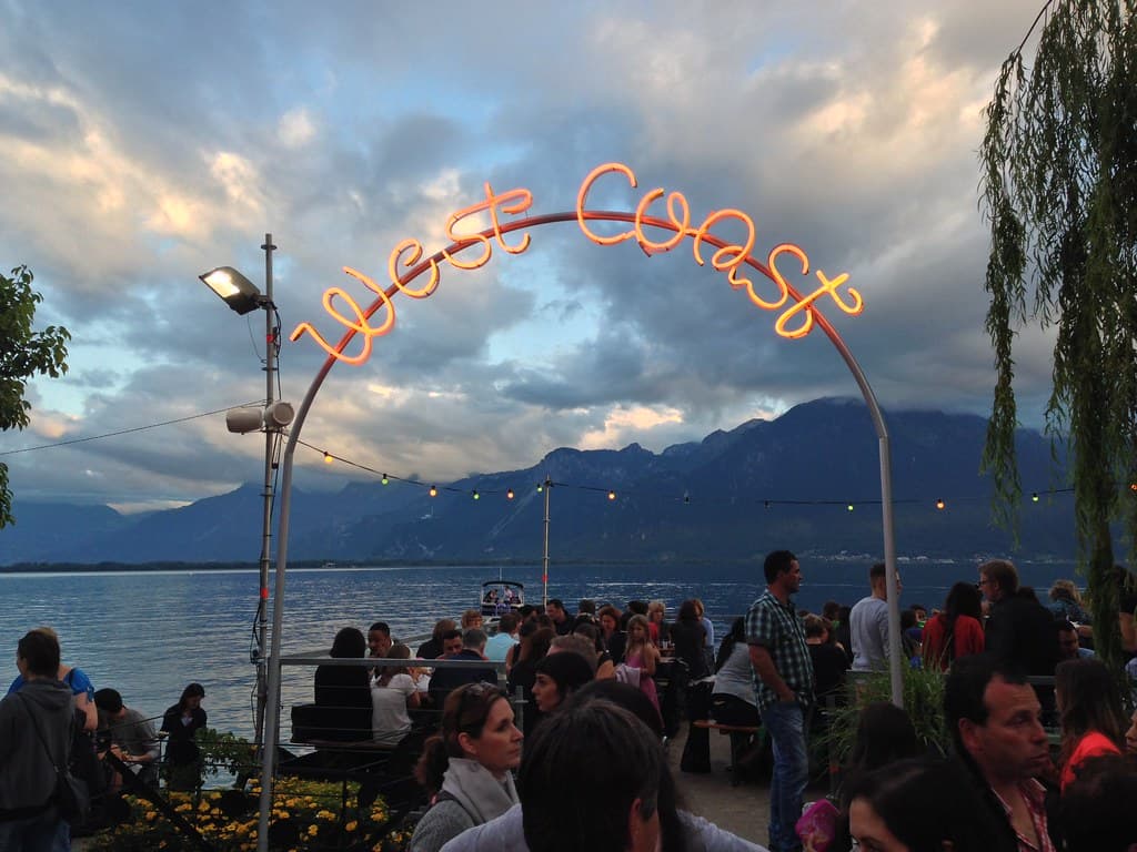 Crowds gather on the lakeside promenade at the Montreux Jazz Festival under an illuminated arch, with Lake Geneva and the mountains behind