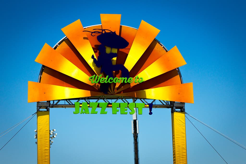 A colourful sunburst arch with a welcome sign stands at the entrance of the New Orleans Jazz and Heritage Festival under a blue sky