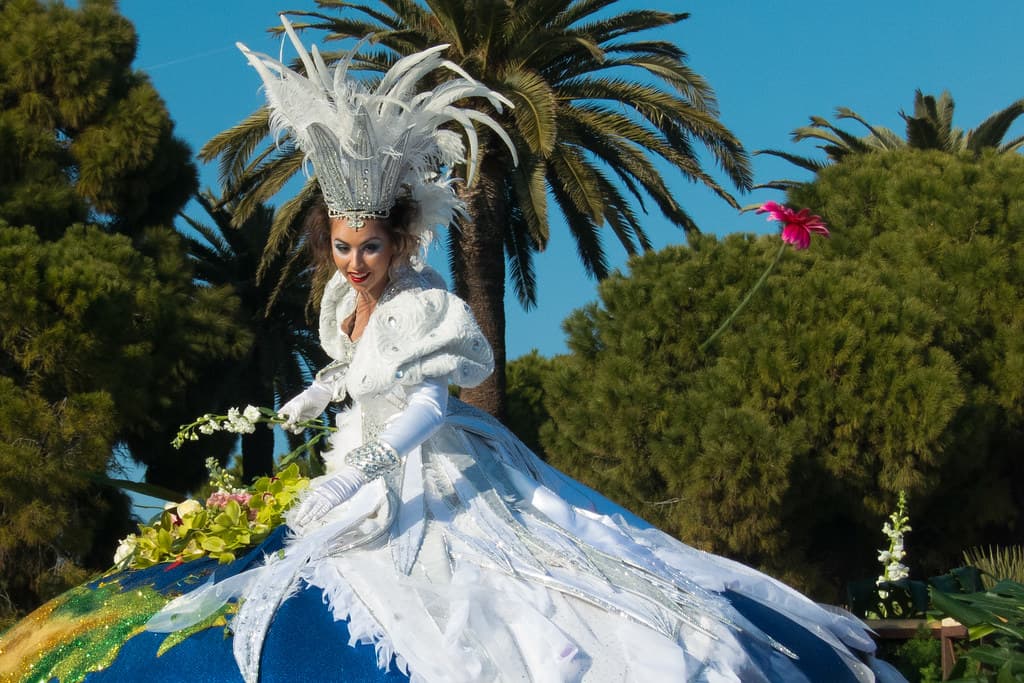 A performer in a white feathered costume rides a flower-covered float under palm trees at the Nice Carnival flower battle