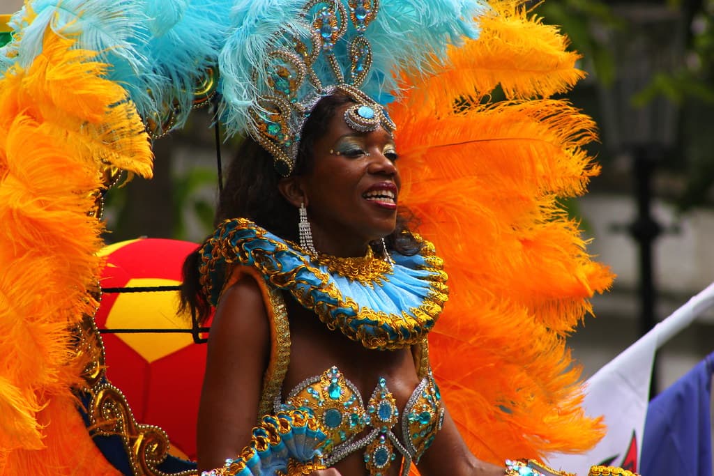 A masquerader in a sequinned costume with huge orange and turquoise feathers dances in the Notting Hill Carnival parade