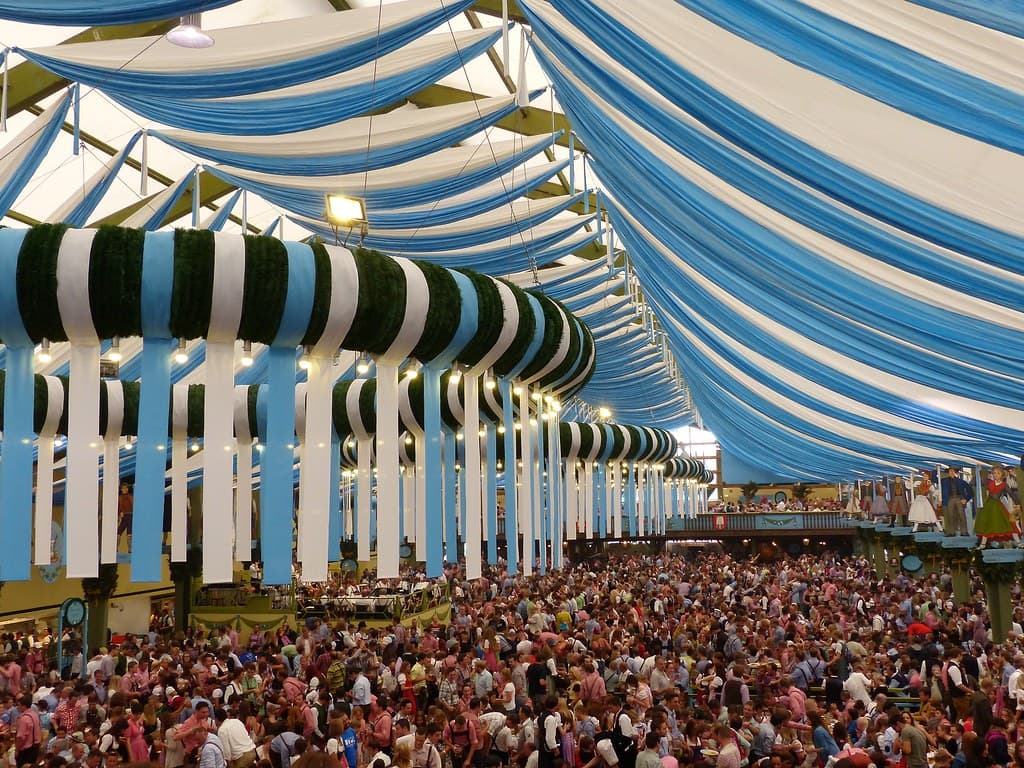 A packed Oktoberfest beer hall under a blue-and-white striped ceiling, with long tables full of drinkers