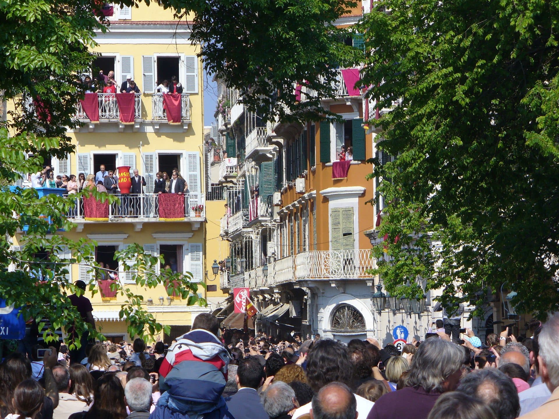 Crowds pack a Corfu Town street beneath balconies hung with red cloths during Orthodox Easter celebrations