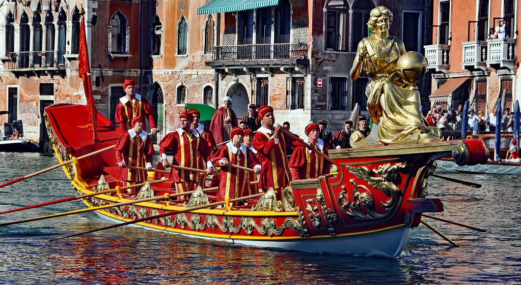 A red and gold ceremonial boat with a gilded statue and rowers in costume glides along the Grand Canal during the Regata Storica