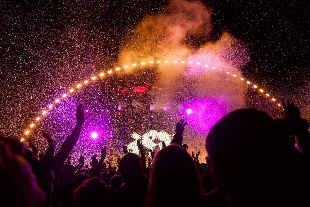 Confetti and coloured lights explode over a crowd with raised hands at a night concert at Roskilde Festival