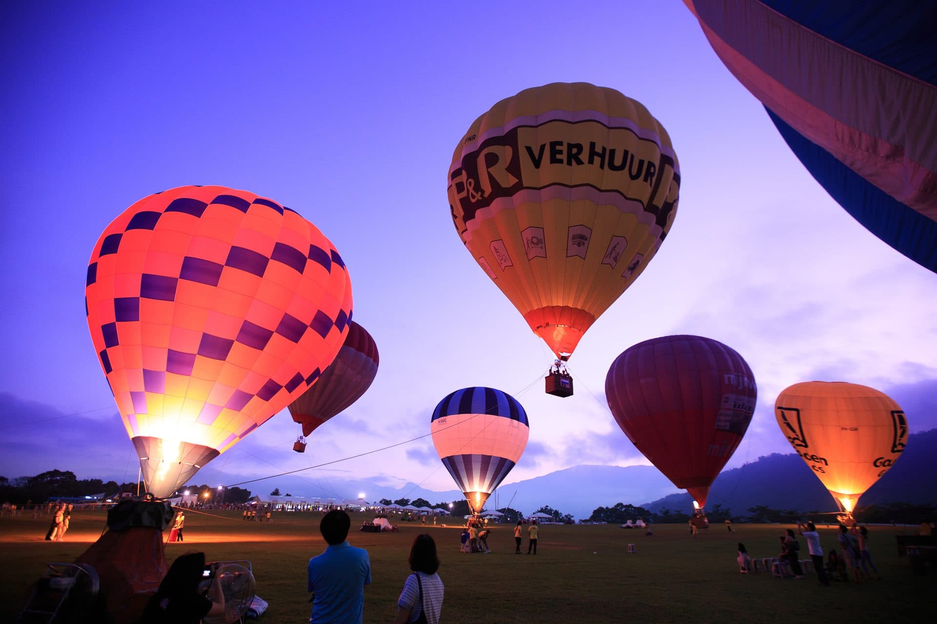 Hot-air balloons glow against a violet dusk sky as visitors watch from the grass at Luye Highland in Taitung