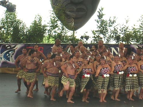 A kapa haka group in traditional dress performs a haka on the stage at Te Matatini, under a large carved mask