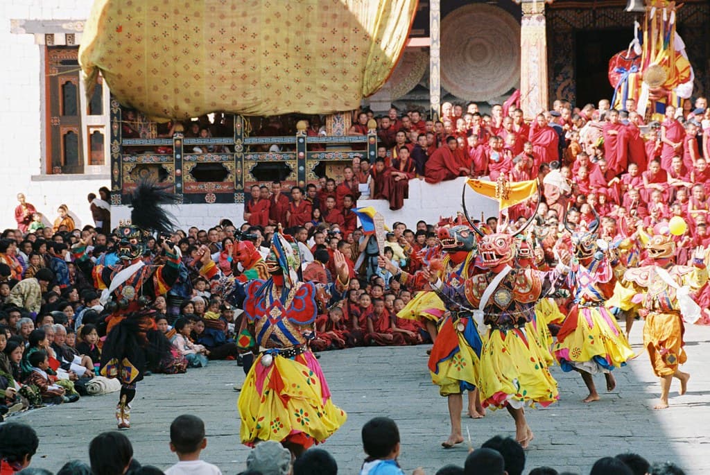 Masked dancers in yellow and red silk skirts spin in the courtyard of Tashichho Dzong during the Thimphu Tshechu, watched by a large crowd