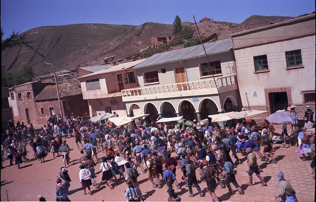 A large crowd of people in traditional dress gathers in the plaza of Macha, Bolivia, under a blue sky during the Tinku festival