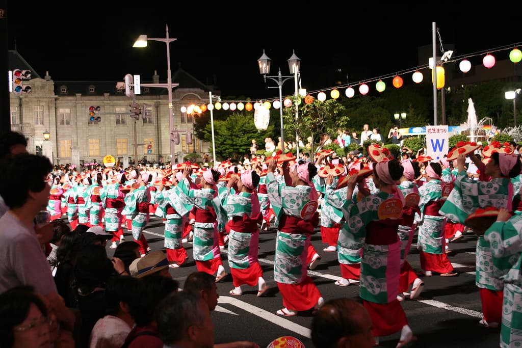 Long lines of dancers in matching yukata twirl flower hats along a lantern-lit street at night in Yamagata