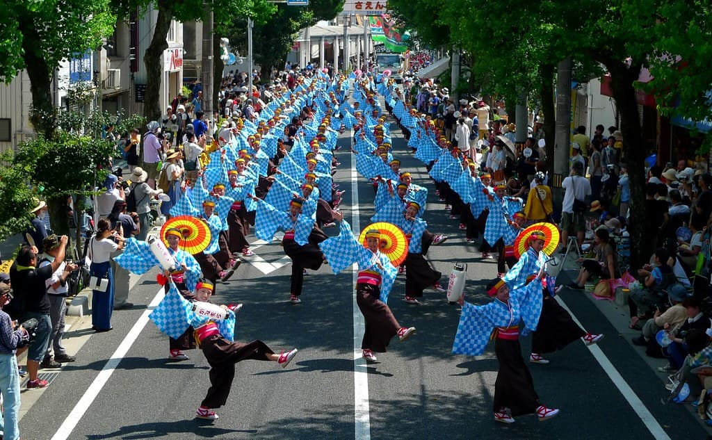 A large yosakoi team in blue costumes dances in formation down a tree-lined street in Kochi, watched by crowds