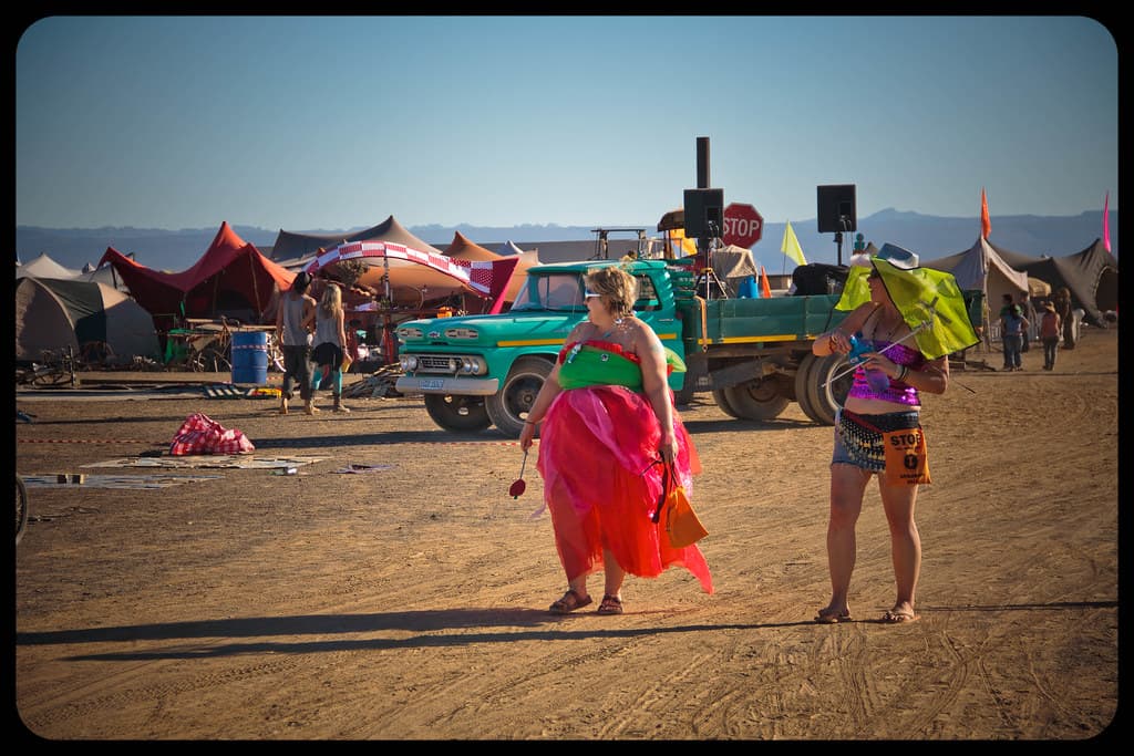 People in colourful costumes walk across the dusty plain of the AfrikaBurn camp, with tents and vehicles behind