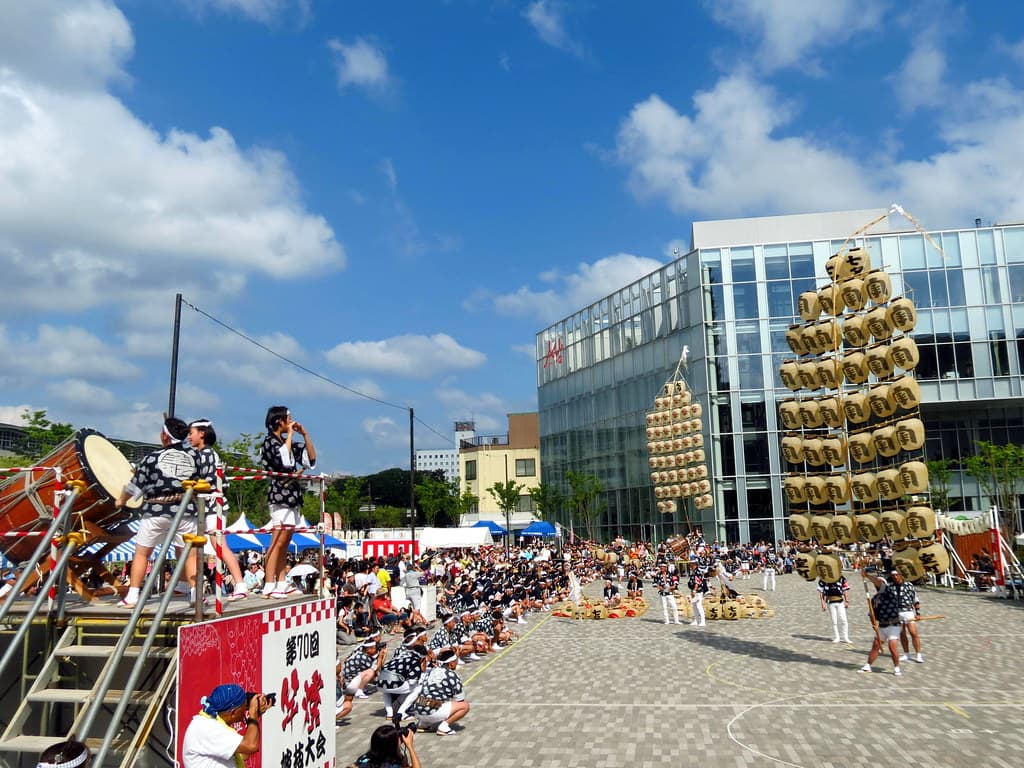 A performer balances a tall kantō pole of white paper lanterns in a city square in daylight, with drummers on a stage and spectators around