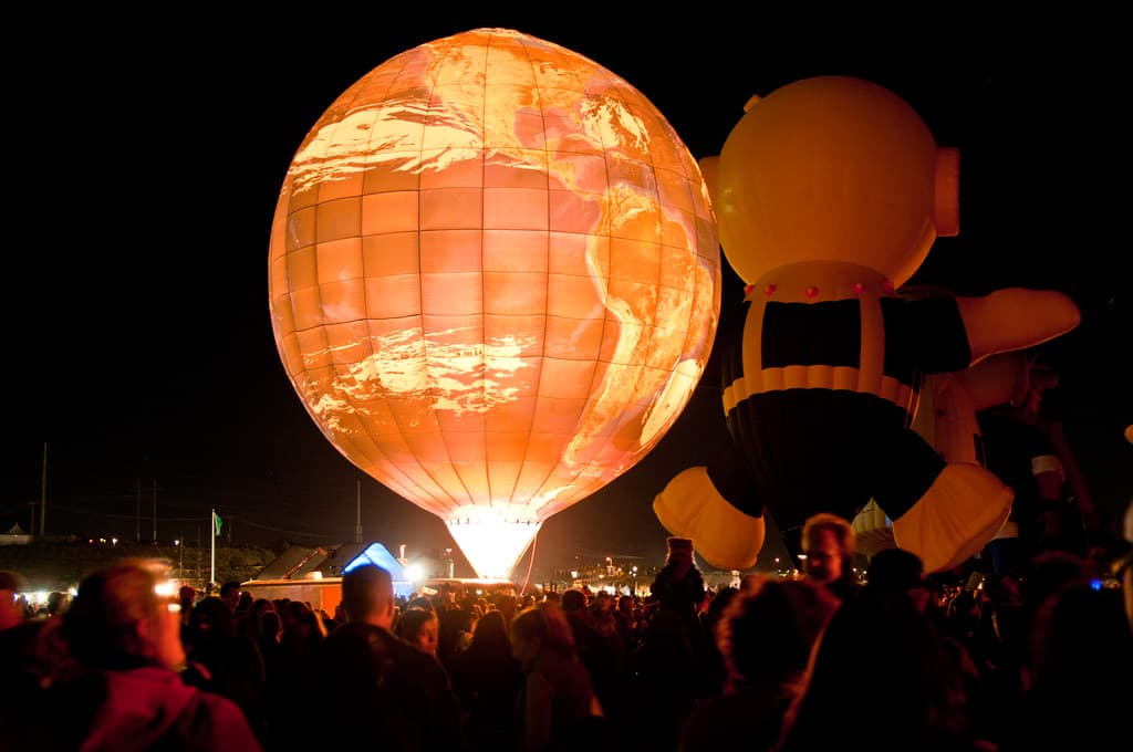 A glowing globe-shaped balloon and a large character balloon are lit up at night in front of a crowd on the Balloon Fiesta field