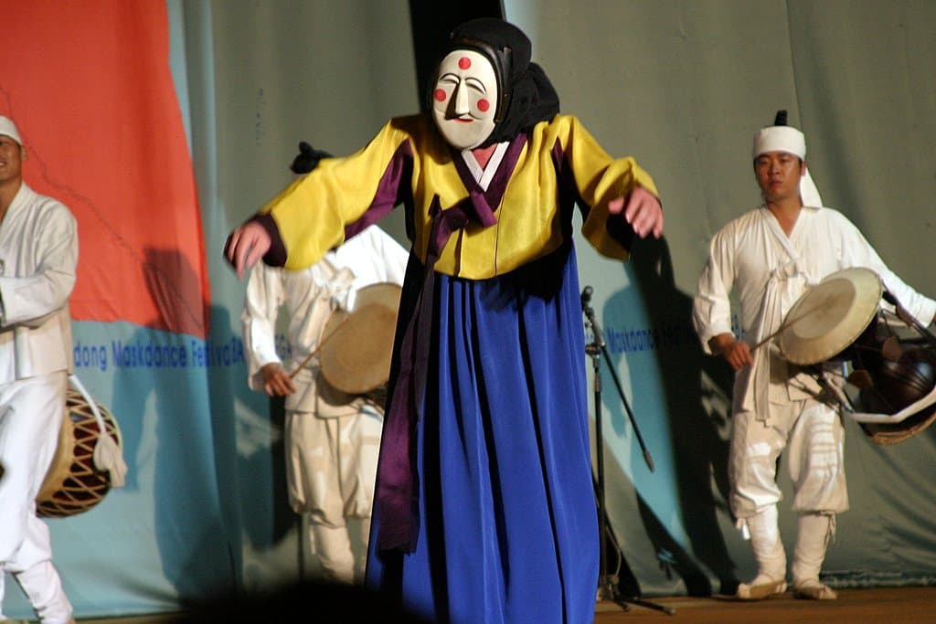 A dancer in a white mask with red dots and a yellow-and-blue hanbok moves in front of two drummers on stage