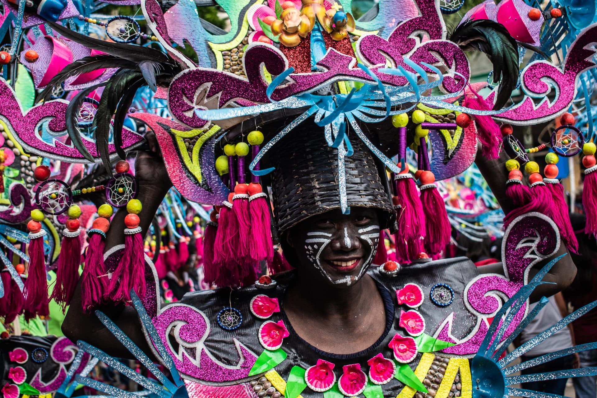 A smiling participant in a glittering costume of magenta and blue foam and feathers, face painted black with white patterns