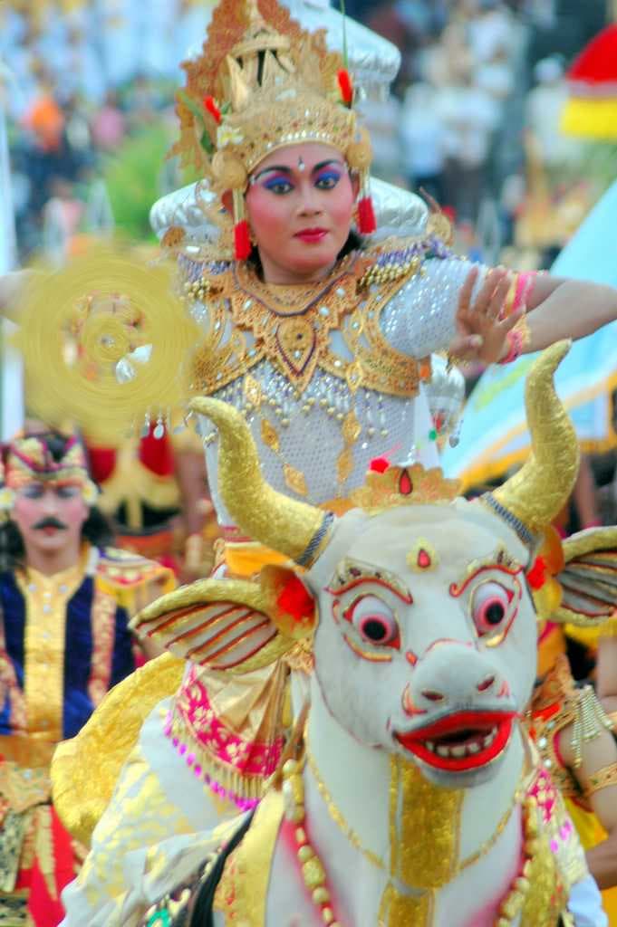 A Balinese dancer in a gold crown and costume rides a decorated white bull figure in a festival procession