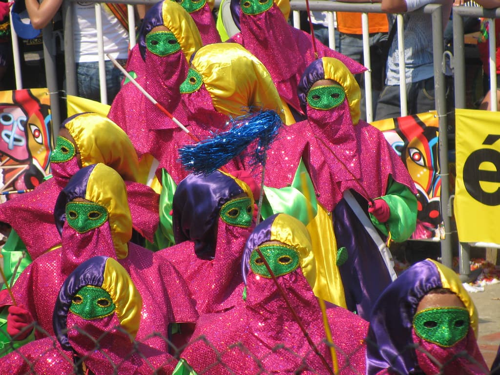 A group of costumed dancers in sequinned pink hoods, purple-and-yellow caps and green masks