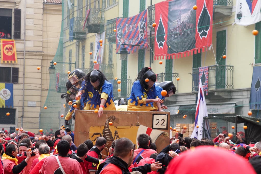 Crowds in red caps fill a square in Ivrea as a cart of masked throwers rolls past during the Battle of the Oranges
