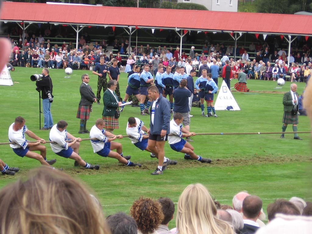 Two tug-of-war teams pull on a rope on the grass at the Braemar Gathering in front of a crowd and the royal pavilion
