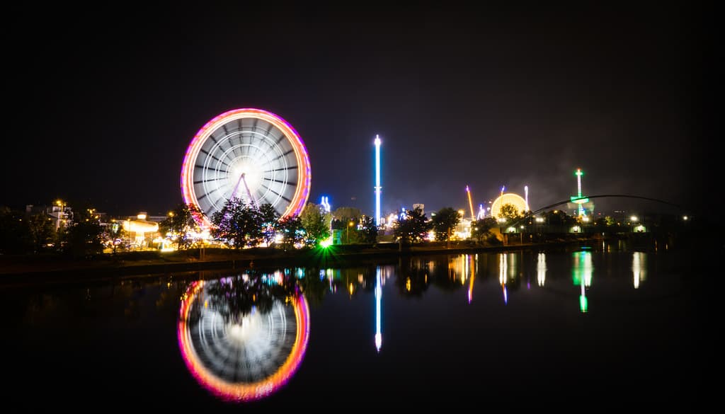 The illuminated Ferris wheel and rides of the Cannstatter Volksfest reflected in the River Neckar at night