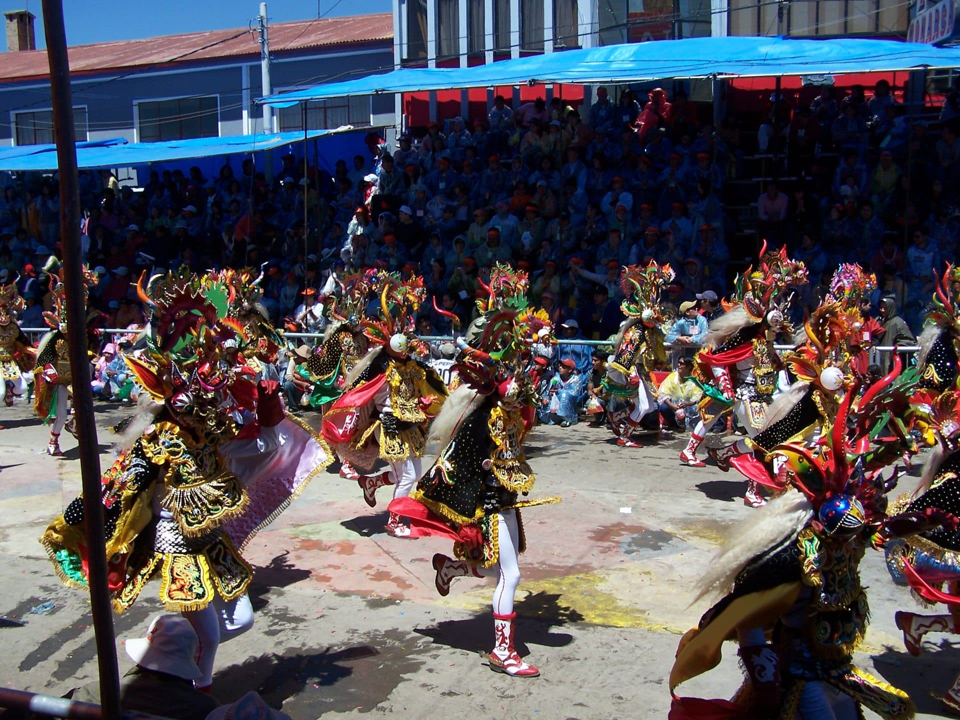 Devil-masked dancers in black and gold costumes with long white mane tails perform on a street in front of crowded stands