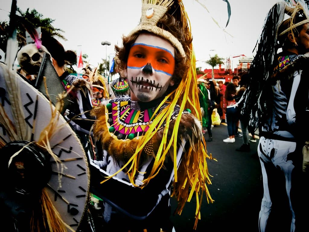 A carnival performer in a painted skull face and a towering feathered costume dances in a Santa Cruz street